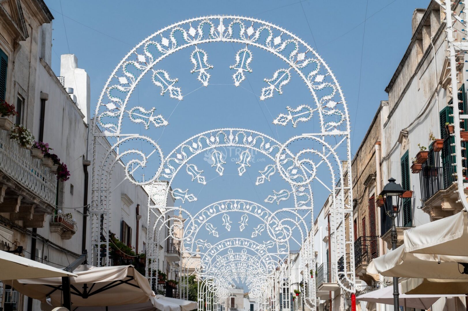 guida-ceglie-messapica-trullionline-19 Via del centro con archi di luminarie bianche, balconi fioriti e ombrelloni sotto il cielo azzurro