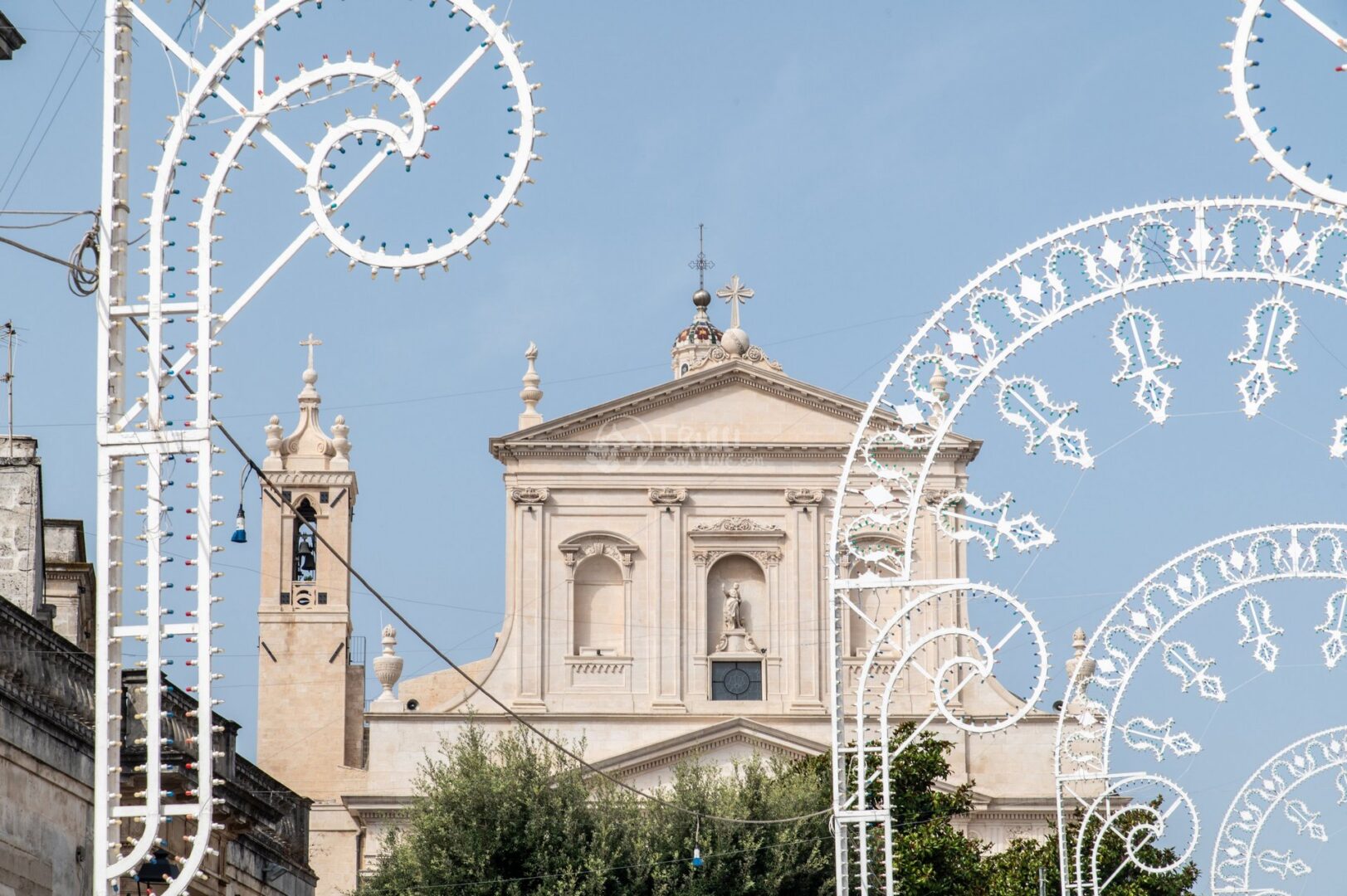guida-ceglie-messapica-trullionline-20 Facciata chiara di una chiesa con frontone e campanile, circondata da archi di luminarie bianche