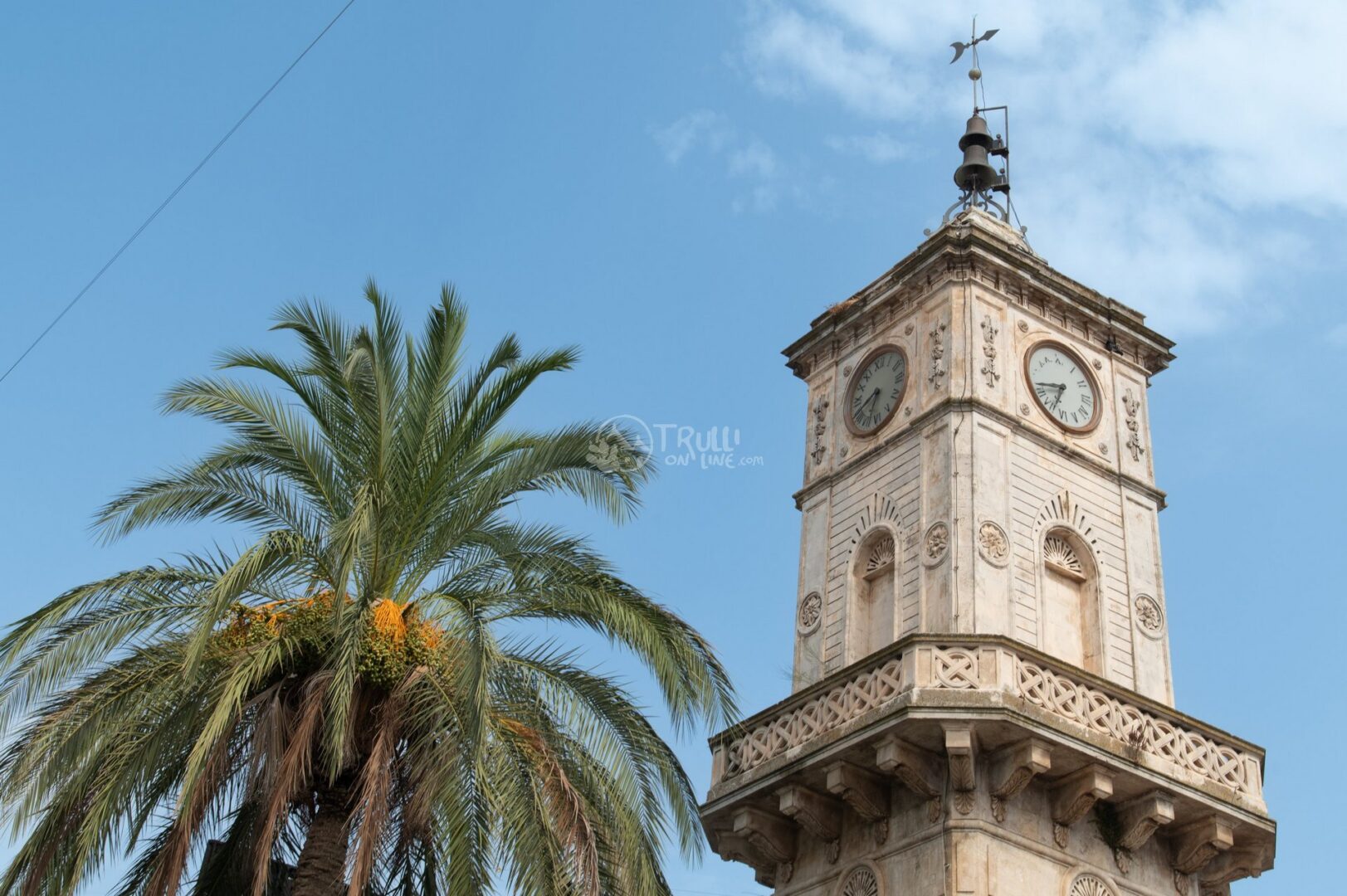 guida-ceglie-messapica-trullionline-21 Torre con due orologi e balconcino in pietra accanto a una palma, sotto un cielo azzurro