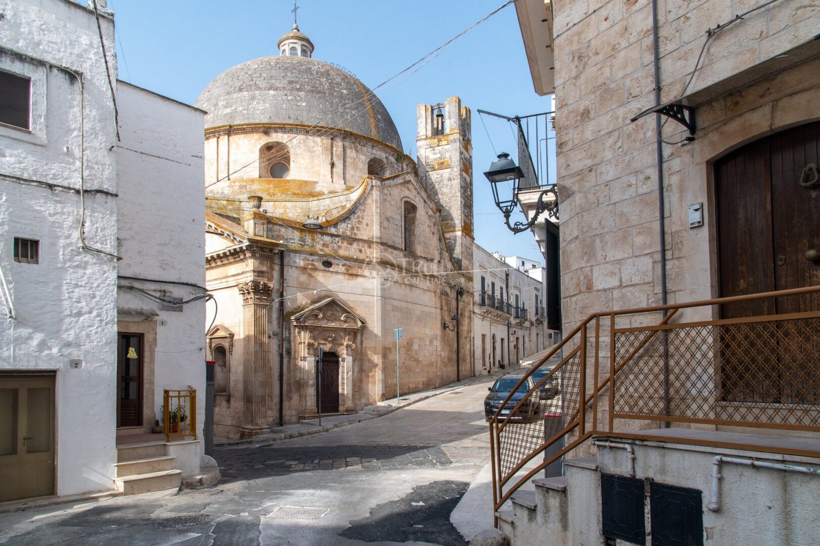 guida-ceglie-messapica-trullionline-26 Una strada del centro con una chiesa dalla cupola e un campanile in fondo, tra case bianche e in pietra