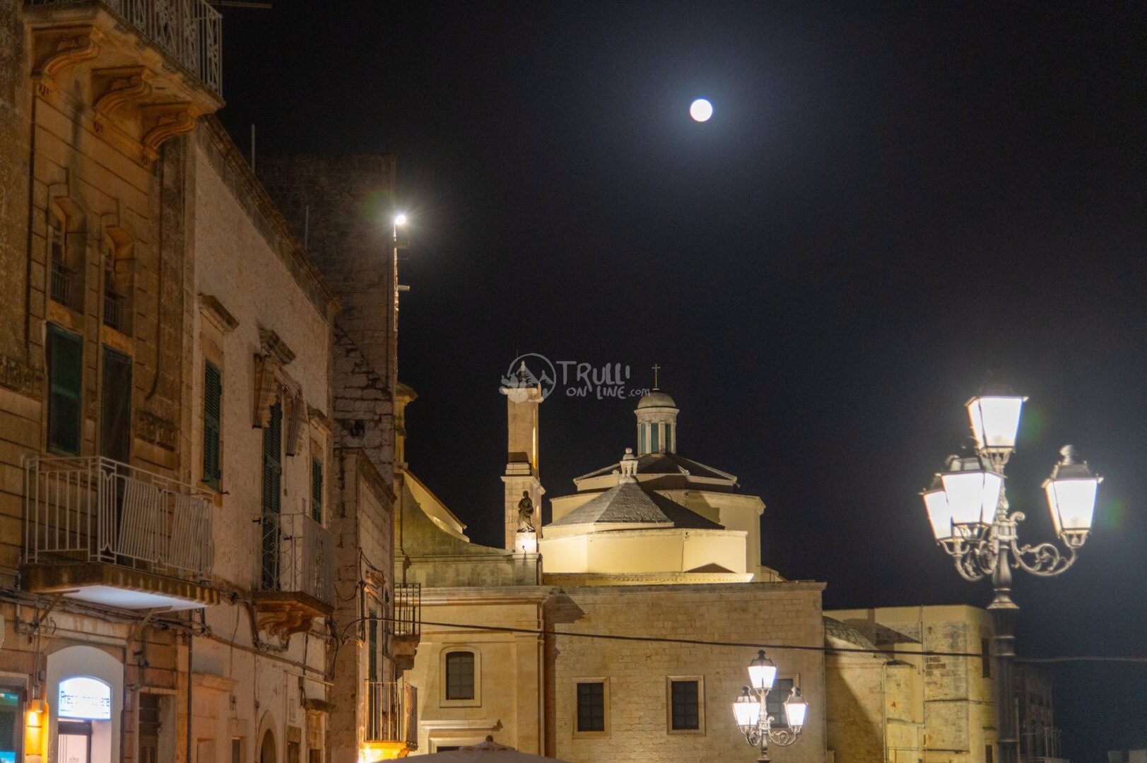 guida-cisternino-trullionline-03 Una chiesa con cupola illuminata di notte accanto a palazzi storici, con luna piena e lampioni
