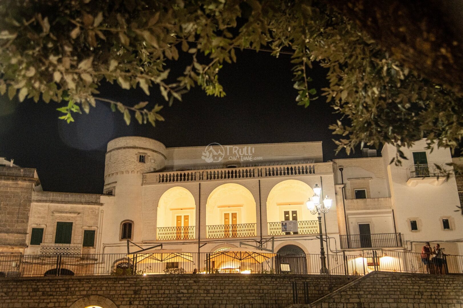 guida-cisternino-trullionline-05 Un palazzo storico bianco con tre arcate illuminate e balaustra, visto di notte tra i rami di un ulivo
