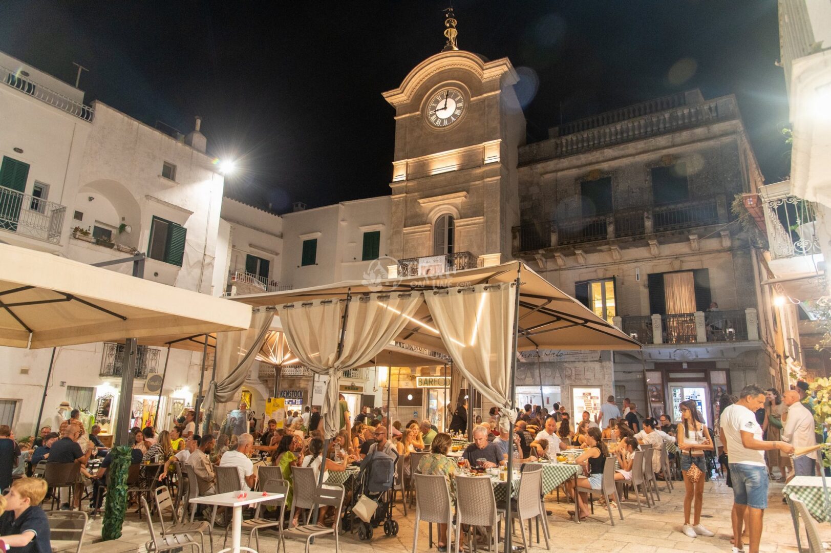 guida-cisternino-trullionline-07 Piazza del centro storico di sera con una torre con orologio e tavoli all'aperto affollati sotto un gazebo