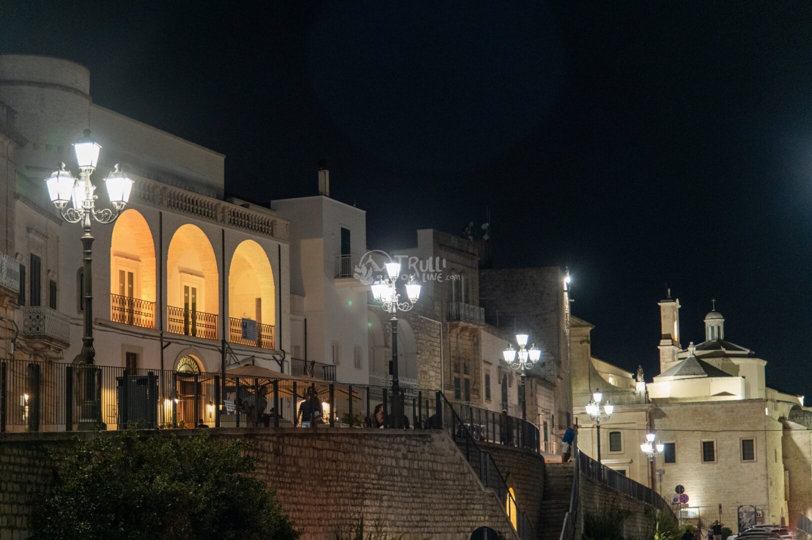 guida-cisternino-trullionline-17 Un palazzo storico con arcate illuminate e una chiesa con cupola, di notte, dietro una scarpata con lampioni