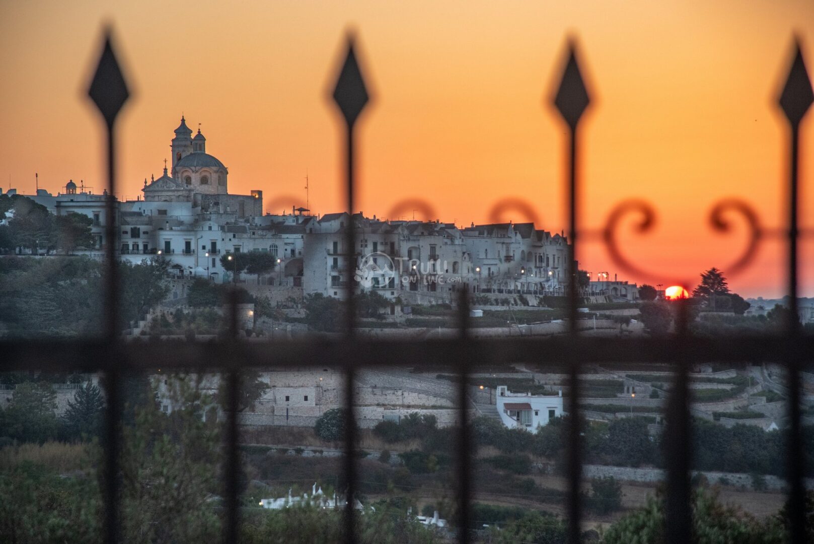 Il paese bianco sulla collina con una chiesa a cupola al tramonto, visto dietro una ringhiera di ferro