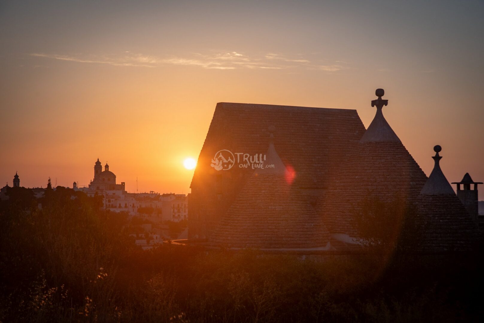 Il sole al tramonto dietro il tetto conico di un trullo, con il paese sullo sfondo