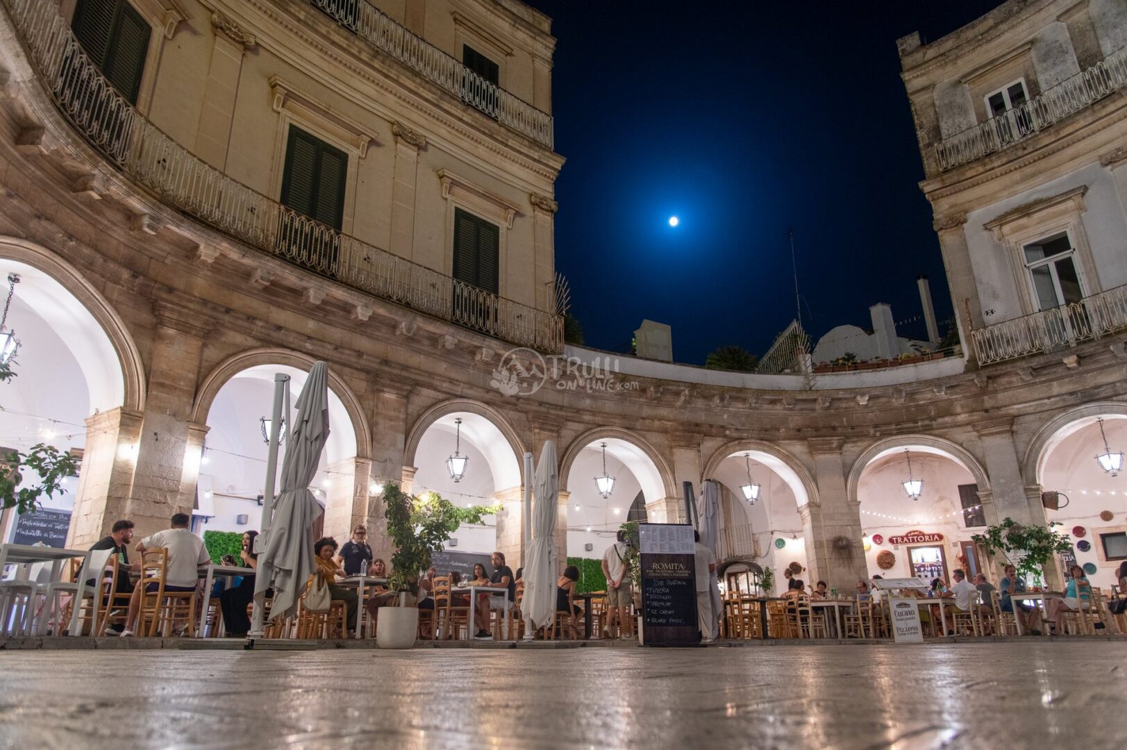 guida-martina-trullionline-07 Piazza semicircolare con portici ad arco, tavoli all'aperto e luna nel cielo notturno a Martina Franca