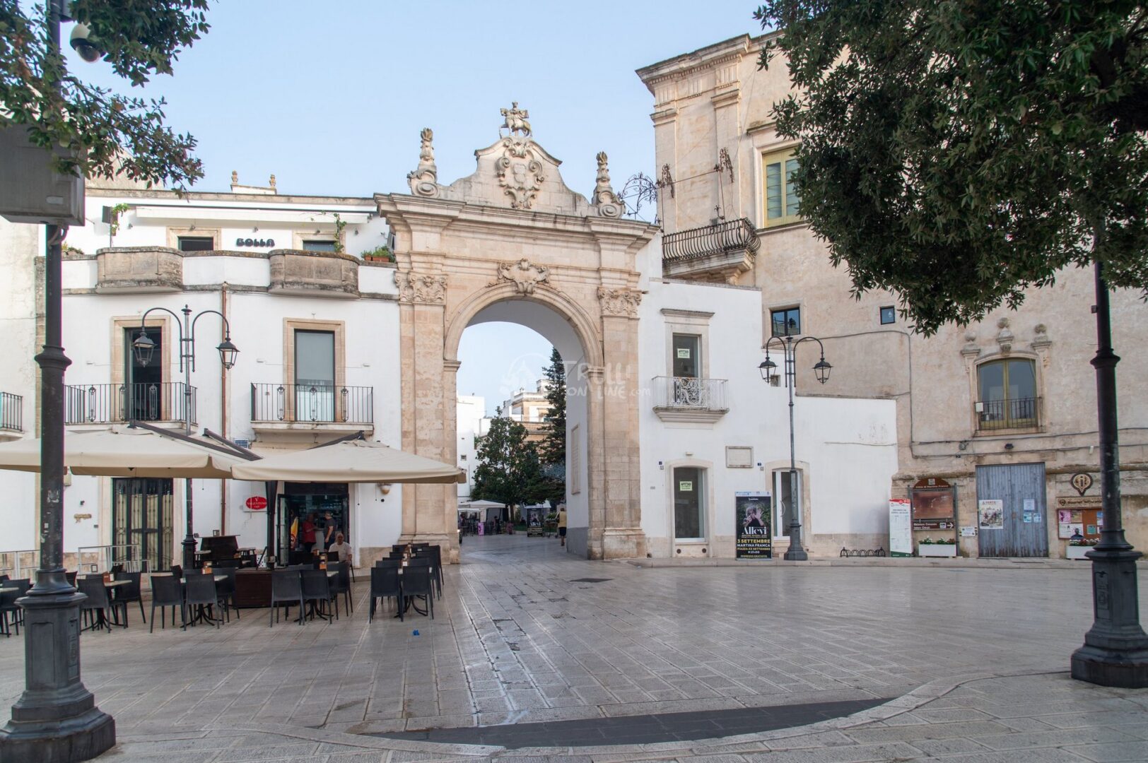 guida-martina-trullionline-09 Arco monumentale barocco in pietra su una piazza con tavolini di un bar, nel centro storico di Martina Franca