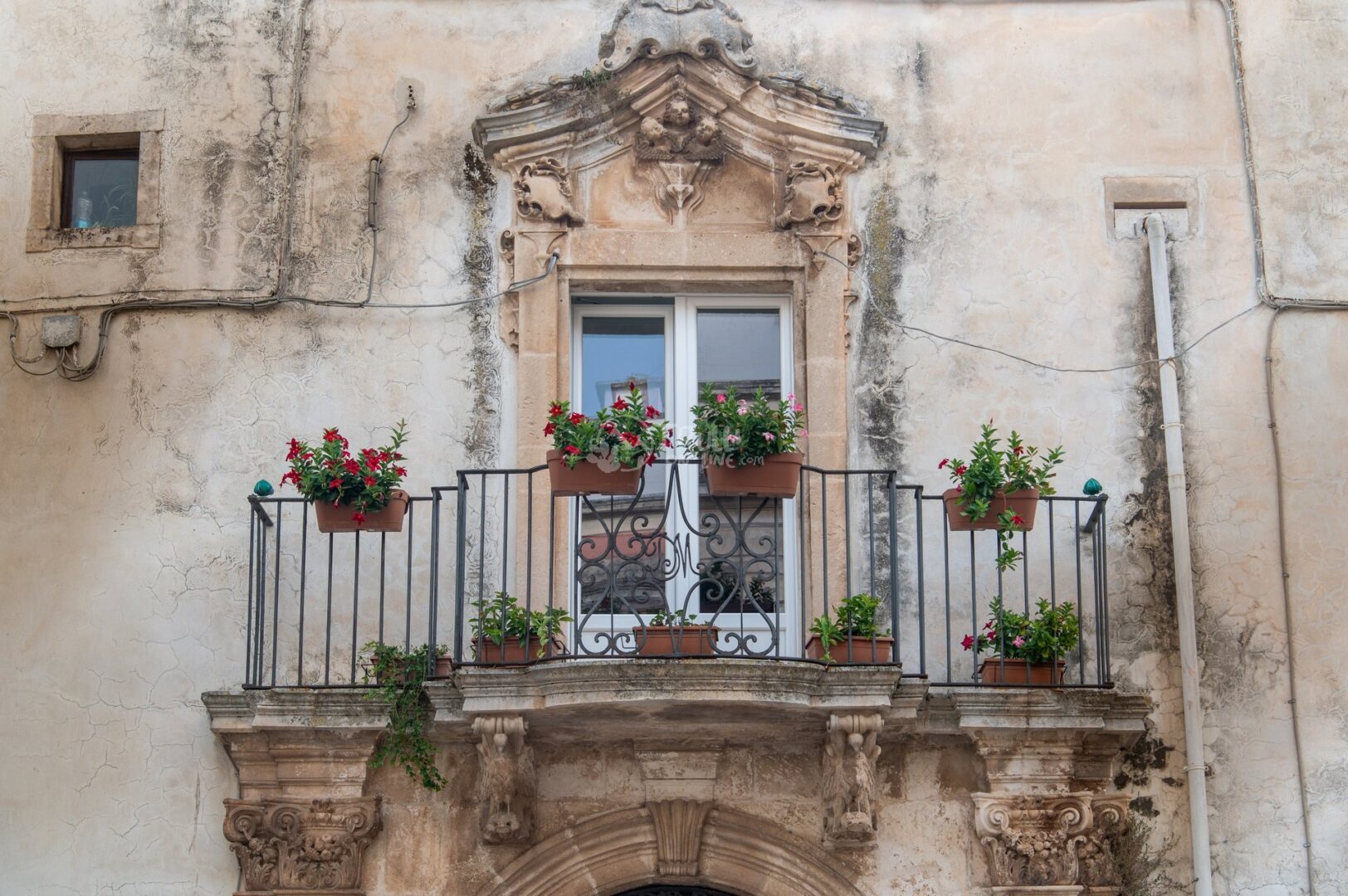 guida-martina-trullionline-14 Balcone fiorito di gerani rossi con ringhiera in ferro e cornice barocca sulla facciata di una casa antica