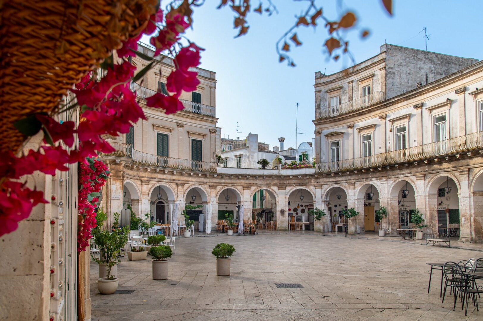 guida-martina-trullionline-18 Piazza porticata semicircolare con vasi di piante e bouganville rosse in primo piano a Martina Franca