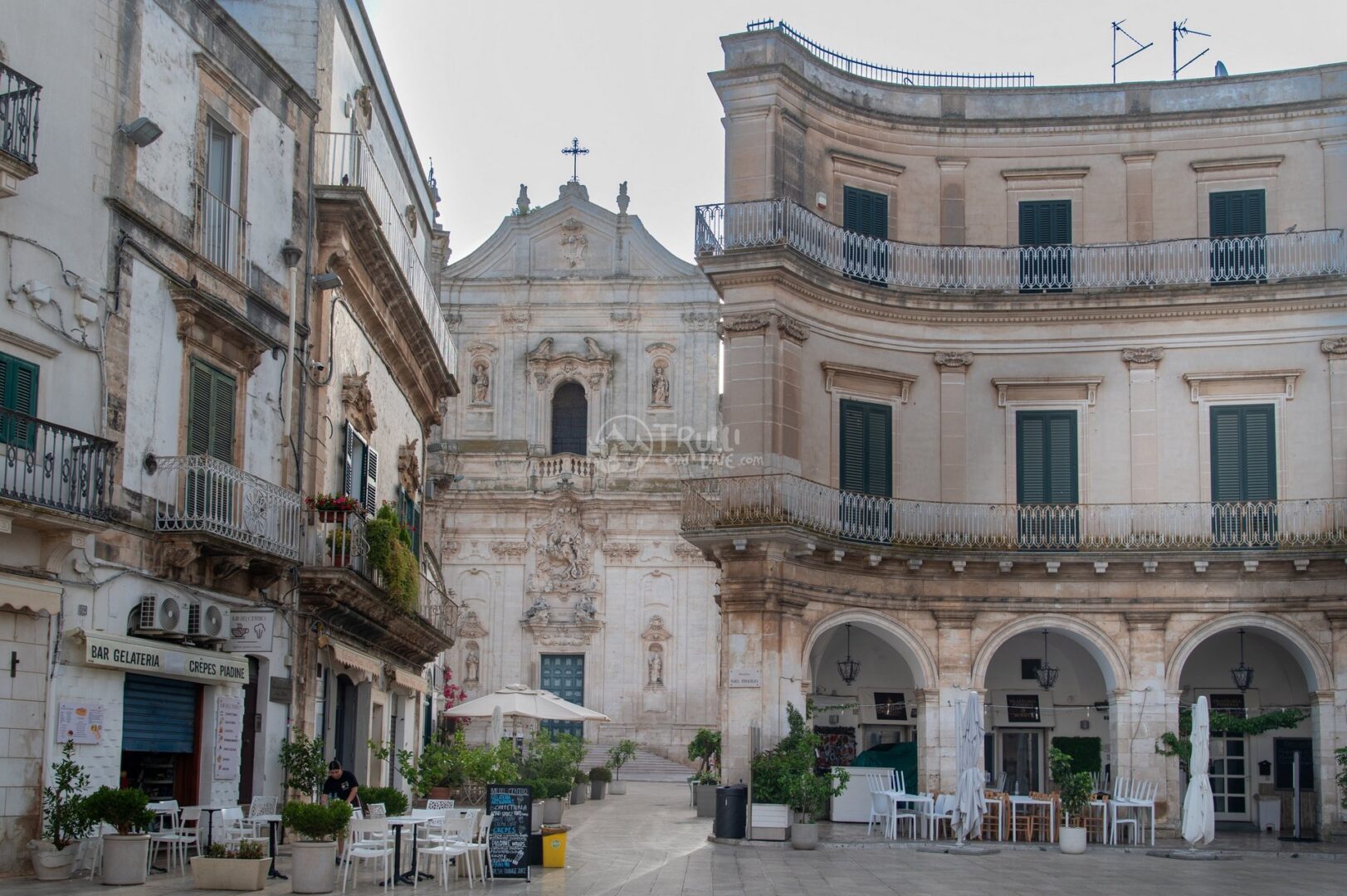 guida-martina-trullionline-19 Piazza con una chiesa barocca sullo sfondo e un palazzo curvo con portici, bar e tavolini a Martina Franca