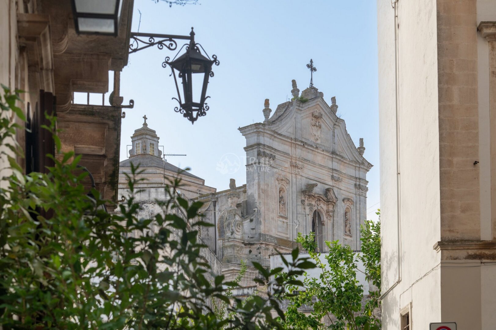 guida-martina-trullionline-22 Facciata di una chiesa barocca con croce tra i tetti, vista da un vicolo con lanterna in ferro e vegetazione