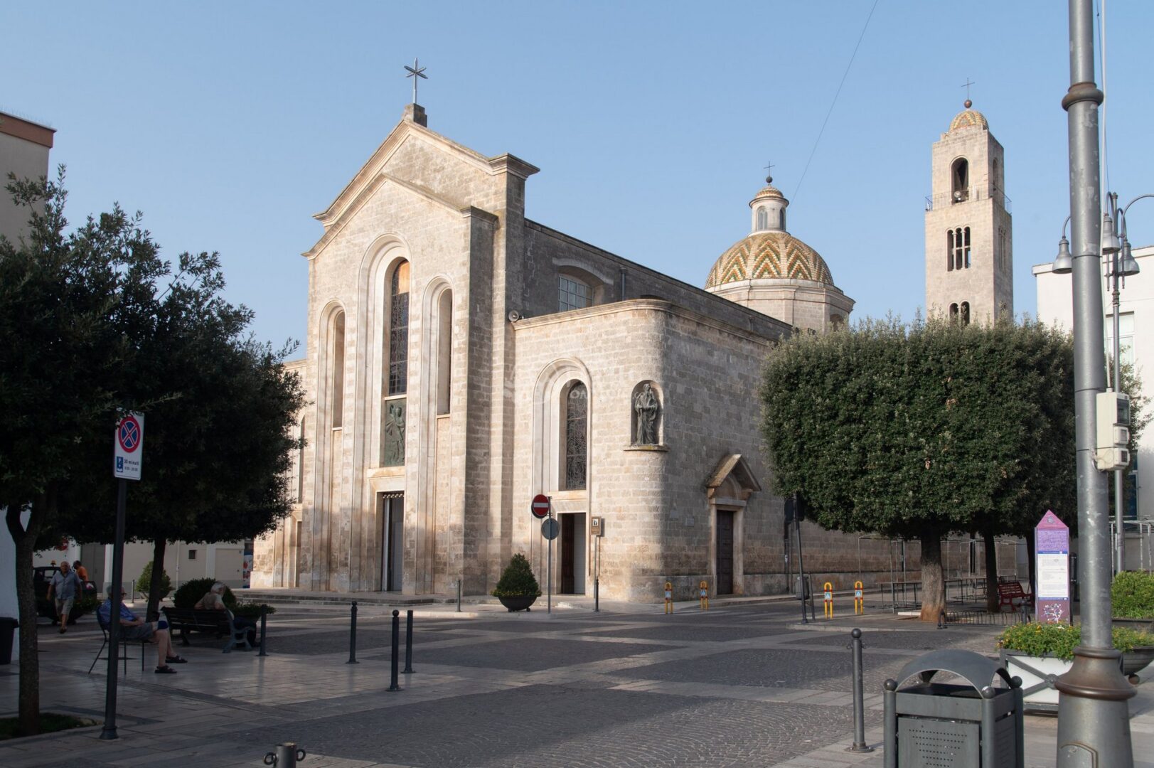 guida-san-michele-trullionline-01 Una chiesa in pietra chiara con cupola decorata e campanile, su una piazza con alberi potati
