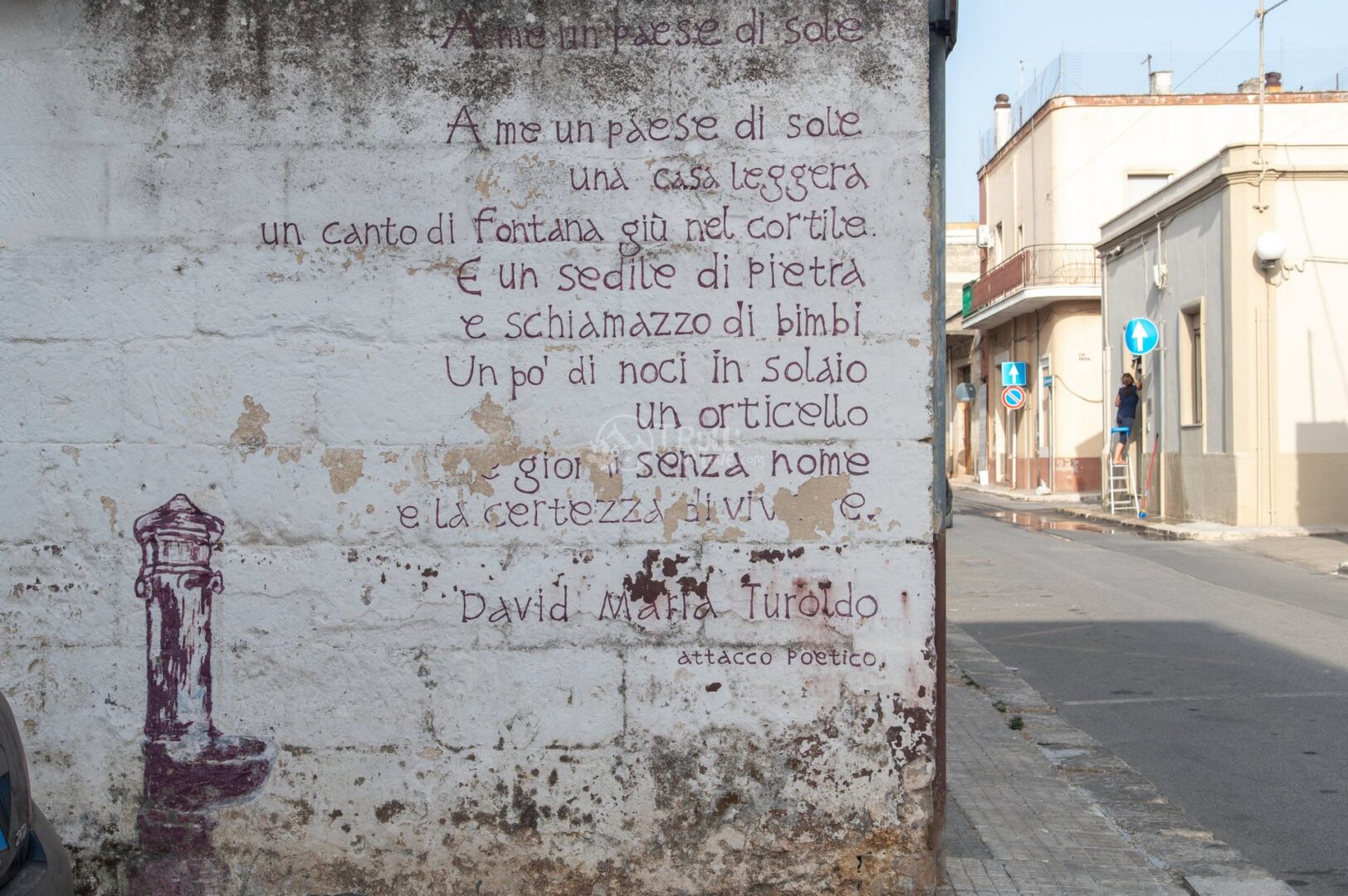 guida-san-michele-trullionline-05 Muro bianco con versi di una poesia dipinti a mano e il disegno di una fontanella, accanto a una strada